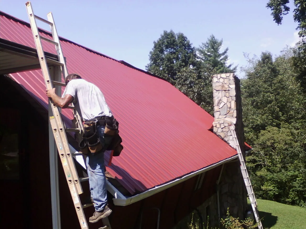Roofer on a ladder beside a red metal roof and stone chimney — Soffit & Fascia Repair by Roofing Brothers in Park Rapids
